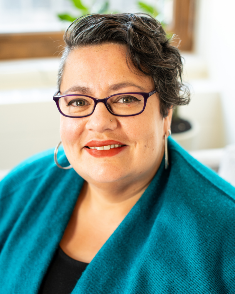 Portrait of a woman with short curly hair and purple glasses wearing a teal shawl, photographed indoors with plants in the background.